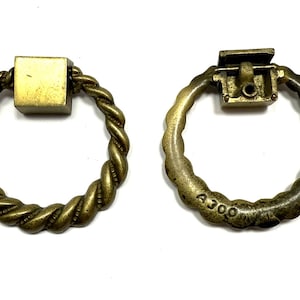 May include: Two antique brass drawer pulls. Each pull features a twisted rope design forming a circular handle. One pull has a square mounting plate, the other has a rectangular plate. The pulls are set against a white background.