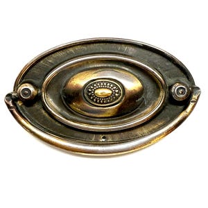May include: Antique brass drawer pull with an oval shape. The center features a smaller oval with a decorative pattern. The pull has a dark, aged finish with hints of gold and silver, and two small circular mounting points.