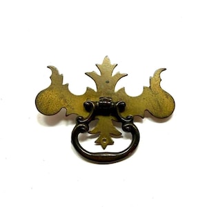 May include: A brass drawer pull featuring a decorative backplate with an ornate, symmetrical silhouette and a central drop handle. The metal surface displays a weathered, aged patina with dark brown accents and intricate, pointed edges.