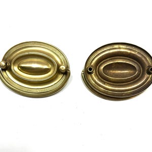 May include: Two antique brass oval drawer pulls. Each pull has a central oval design, surrounded by a detailed border. One pull has a handle, while the other has two holes for attachment. The pulls are a warm, aged brass color.