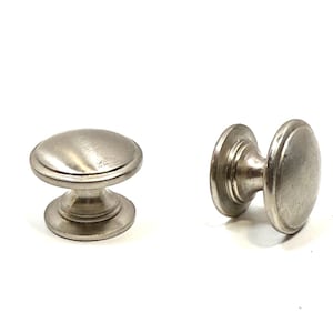 May include: Two silver-toned metal cabinet knobs with a smooth, rounded top surface and a tiered, circular base. The knobs feature a polished finish with subtle metallic reflections, showcasing a simple, functional design for furniture hardware.