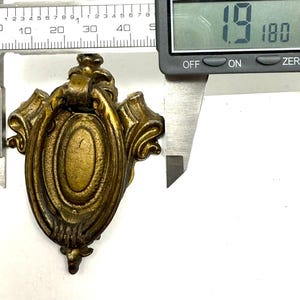 May include: Antique brass-toned furniture pull with an oval center and ornate scrollwork. The pull measures approximately 1.9 inches in length. A digital caliper is used to measure the pull.