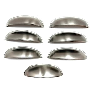 May include: Seven brushed nickel cup pulls arranged on a white background. The pulls have a half-moon shape with a smooth, metallic finish. These cabinet hardware pieces are ideal for kitchen or bathroom renovations.