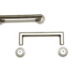 May include: Two antique-style cabinet pulls and two matching circular backplates. The pulls are rectangular and have a distressed, silver-gray finish. The backplates have a concentric circle design. The hardware is for cabinets and drawers.