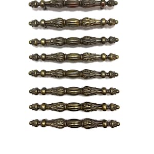 May include: Eight antique bronze-colored drawer pulls with ornate detailing. Each pull features a central, ribbed section flanked by decorative ends. The pulls are arranged horizontally against a white background, showcasing their aged finish and intricate design.