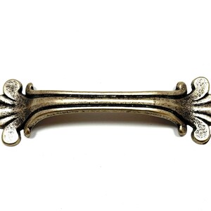 May include: Antique-style cabinet pull with a fleur-de-lis design. The handle is a muted gold color with a distressed finish, and is approximately 5 inches long. The decorative ends feature a detailed floral pattern.