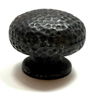 May include: A dark, hammered metal cabinet knob with a rounded top and a short, cylindrical base. The knob has a textured surface and a dark, aged appearance, suggesting a rustic or vintage style. The knob is designed for use on drawers or cabinet doors.
