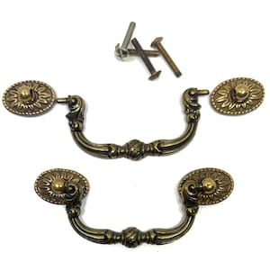 May include: Two antique brass-toned metal drawer pulls with swinging handles and oval backplates featuring a sunburst pattern. The set includes four matching metal mounting screws for installation, showing a dark metallic finish and ornate detailing.