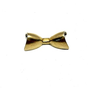 May include: A gold-colored bow tie pin with a slightly aged appearance. The pin is made of metal and has a classic bow tie shape. The bow tie is centered on a white background.