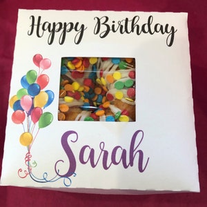 May include: White gift box with a window showing a donut cake with white frosting and colorful sprinkles. The box has the text "Happy Birthday Sarah" printed on it with colorful balloons.