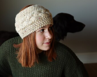Può includere: Una donna con i capelli rossi indossa una fascia per capelli in maglia color crema con un motivo a treccia. Indossa un maglione verde scuro.