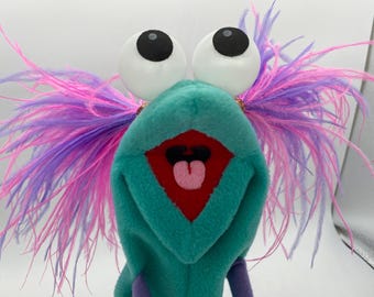 May include: A teal plush hand puppet featuring large white googly eyes with black pupils, a red mouth interior, a pink tongue, and vibrant pink and purple feather hair extending from the sides of the head. The puppet has a soft fabric texture.