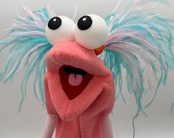 May include: A pink hand puppet with large white spherical eyes featuring black pupils. The puppet has a wide open mouth with a red interior and a small pink tongue. It is adorned with long, wispy turquoise and white feathers on both sides.