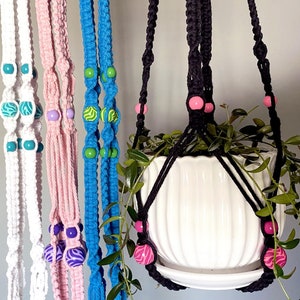 以下が含まれることがあります： 異なる色のコードとビーズが付いた、5つのマクラメ製のプラントハンガー。ハンガーは、白、ピンク、青、黒、緑など、さまざまな色で作られています。ハンガーはカラフルなビーズで飾られています。ハンガーの1つは、緑の植物が入った白いセラミック製の鉢を持っています。