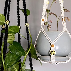 May include: Two macrame plant hangers with yellow and brown patterned beads. One hanger is black and the other is white. A green plant is hanging in the black hanger.