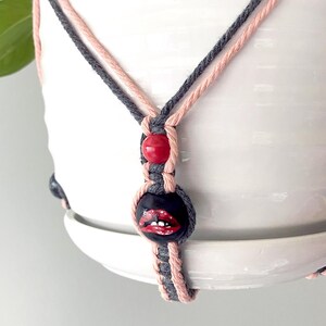 Op de afbeelding: Een macramé plantenbak in grijs en roze, met een witte keramische pot met groene bladeren. De hanger heeft een zwarte kraal met rode lippen en een rode kraal.