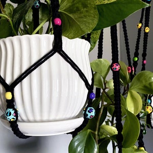 May include: Black macrame plant hanger with colorful beads. The plant hanger holds a white ceramic pot with a green plant. The beads are various colors, including pink, yellow, blue, and leopard print. The plant hanger is suspended from the ceiling.