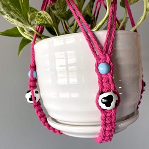 Könnte beinhalten: Ein rosa Makramee-Pflanzenhänger mit schwarzen und weißen Kuhflecken-Perlen und hellblauen Perlen. Der Hänger hält einen weißen Keramiktopf mit einer Pflanze.