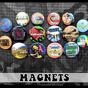 May include: A collection of 15 round magnets featuring various band logos and album covers. The magnets are colorful and feature a variety of designs, including a dinosaur, a cat, a microphone, and a band playing on a stage. The magnets are arranged in a circle on a gray background. The word "MAGNETS" is printed in bold black letters below the magnets.