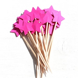 Könnte beinhalten: Ein Bündel rosa Papiersterne auf Holzspießen. Die Sterne sind in einem lockeren Cluster angeordnet, wobei die Spitzen der Sterne nach außen zeigen.