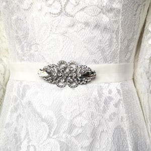 Könnte beinhalten: Weißes Spitzenkleid mit Satinband und funkelndem Strass- und silberfarbenem Metallbesatz. Das Kleid hat lange Ärmel und ein detailliertes florales Spitzenmuster. Der Gürtel befindet sich in der Taille.