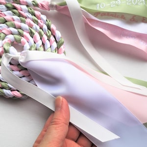 May include: A braided rope of white, pink, and green satin ribbon with four separate ribbons attached. The ribbons are white, pink, light pink, and green. The green ribbon has the text "10-29-2021 Patrick & Erin" embroidered on it. The light pink ribbon has the text "I love you" embroidered on it.