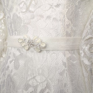 Könnte beinhalten: Nahaufnahme eines weißen Spitzenkleides mit dekorativem Gürtel. Das Kleid weist komplizierte florale Spitzmuster auf und ist mit kleinen Perlen verziert. Der Gürtel hat eine dekorative Blumenschnalle mit Strasssteinen, die Eleganz verleiht.