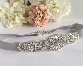 May include: A silver satin bridal sash with a crystal embellishment. The embellishment features a floral design with clear rhinestones.