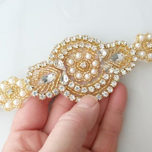 May include: An elaborate gold-coloured embellishment with a floral design. It features clear rhinestones, small pearls, and gold beads. The piece is approximately 15 cm long.