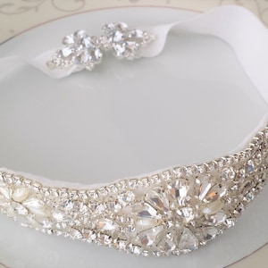 Könnte beinhalten: Ein weißes Stirnband mit einem funkelnden Kristall- und Strassdesign. Das Stirnband hat ein dekoratives Band aus klaren Kristallen und Strasssteinen mit einem floralen Akzent in der Mitte. Das Band ist etwa 2,5 cm breit.