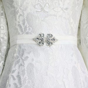 Könnte beinhalten: Weißes Spitzenkleid mit einem weißen Satinband und einem Kristallbesatz.