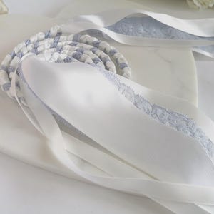 Puede incluir: Una cinta trenzada blanca y azul con cintas de satén blancas. Las cintas de satén tienen un delicado ribete de encaje azul. La cinta trenzada está enrollada y las cintas de satén fluyen.