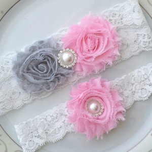 Op de afbeelding: Twee witte kanten kousenbandjes met een roze en grijze stoffen bloemenversiering. Elke bloem heeft een parel in het midden en een strass accent.