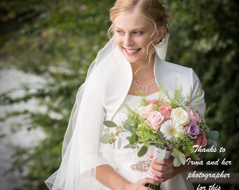 May include: A bride in a white dress and veil holds a bouquet of pink, white, and purple flowers. The bouquet is wrapped in white lace and tied with a pink ribbon. The bride is smiling and looking to the side. The text "Thanks to Irma and her photographer for this beautiful pic!" is in the lower right corner of the image.