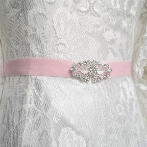 Könnte beinhalten: Nahaufnahme eines weißen Spitzenkleides mit einem hellrosa Schärpe. Die Schärpe ist mit einer glitzernden silbernen und klaren Kristallverzierung versehen. Das Kleid hat aufwendige Spitzendetails und ein zartes Design.