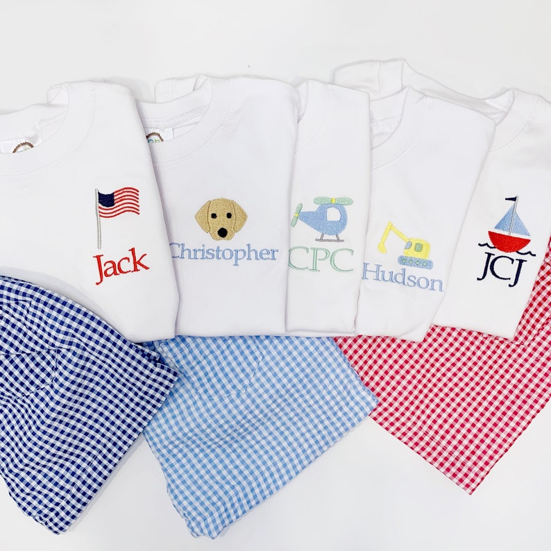 May include: Five white t-shirts with embroidered designs and names, including an American flag, a dog, a helicopter, a construction vehicle, and a sailboat. Each shirt is paired with a checkered pattern short.