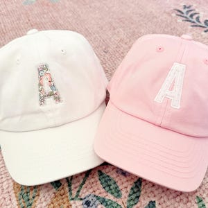 May include: Two baseball caps, one white and one pink, both with the letter 'A' embroidered on the front. The white cap has a floral pattern on the 'A', while the pink cap has a white 'A'.
