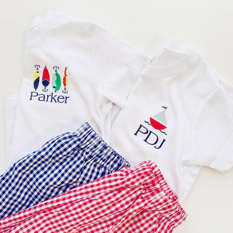 May include: Two white t-shirts with embroidered designs. One shirt has a sailboat with the initials "PDJ" and the other has a row of fishing lures with the name "Parker". Two pairs of gingham shorts, one blue and white and one red and white.