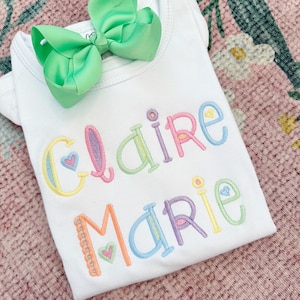 May include: A white onesie with the name "Claire Marie" embroidered in rainbow colors with hearts. The onesie has a green bow on top.