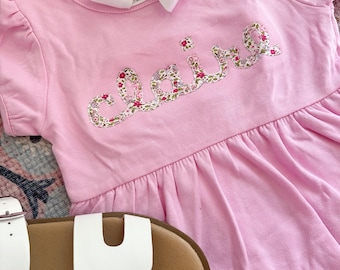 May include: A pink baby dress with short sleeves and a floral print name "claire" on the front.