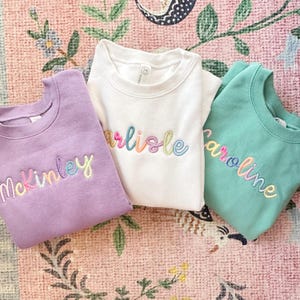 May include: Three crewneck sweatshirts in lavender, white, and teal. Each sweatshirt has a name embroidered in rainbow thread: McKinley, Carlisle, and Caroline. The sweatshirts are folded and arranged on a patterned pink and green surface.