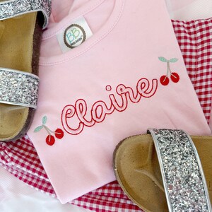 May include: Pink t-shirt with the name "Claire" embroidered in red cursive with two red cherries on each side. The shirt is on top of a red and white gingham fabric.