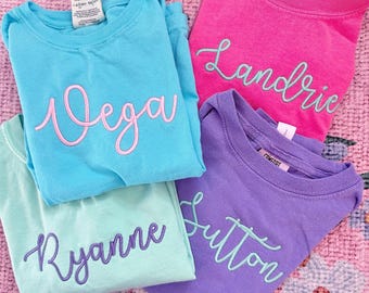 May include: Four t-shirts with different names embroidered on them in a cursive font. The shirts are in various colors, including light blue, pink, purple, and green.
