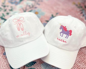 May include: Two white baseball caps with embroidered designs. One cap features a pink ballerina shoe with a bow and the name "molly" embroidered on the front. The other cap features a purple unicorn with a pink mane and the name "reese" embroidered on the front.