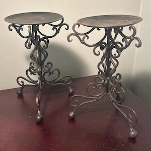 May include: Two ornate, dark metal candle holders. Each has a round top, a decorative, swirling stem, and three ball-footed legs. The candle holders are on a dark surface.