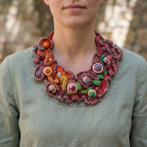 Può includere: Una colorata collana all'uncinetto fatta a mano con bottoni decorativi. La collana presenta sfumature di arancione, rosso, verde e viola. I bottoni hanno disegni floreali e fantasia. Un accessorio artistico unico.