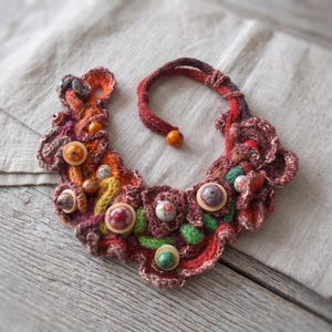 May include: A crochet necklace featuring a vibrant, multi-coloured yarn base in shades of red, orange, yellow, and green. The piece is embellished with various round, patterned buttons and small wooden beads, with a single bead closure at the end.