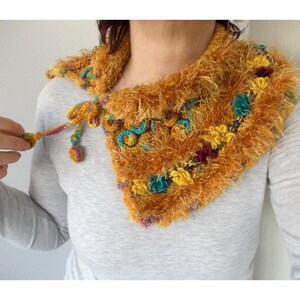 Op de afbeelding: Een handgemaakte, gehaakte nekwarmer in tinten goud, blauwgroen en paars. De nekwarmer heeft een pluizige textuur en is versierd met kleine, kleurrijke bloemaccenten. Een striksluiting is zichtbaar.
