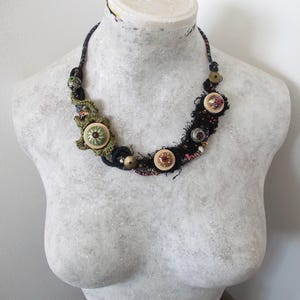 Op de afbeelding: Een ketting met een donker stoffen koord versierd met diverse ronde knopen in crème, groene en metallic tinten, afgewisseld met getextureerd zwart garen, kleine kralen en groene gehaakte accenten in een asymmetrisch ontwerp.