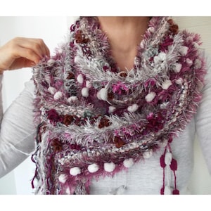 May include: A handmade, fluffy scarf in shades of purple, white, and grey. The scarf features a textured design with white pom-poms and fringe detailing. The scarf is worn around the neck.